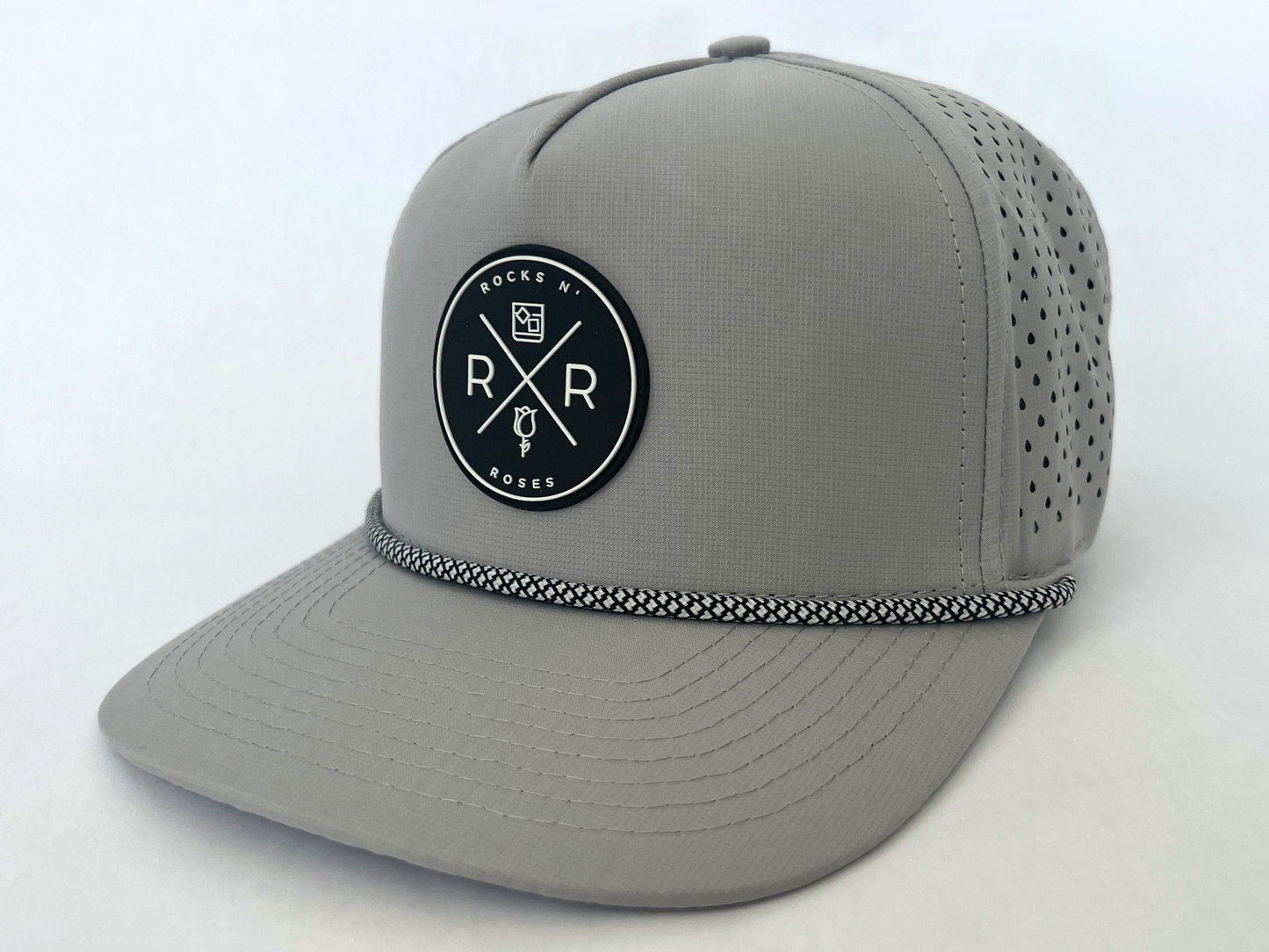 Close up shot of the Rocks N' Roses Gray Performance Golf Hat. This 100% polyester snapback hat is water resistant, offers some stretch and has that new school flat brim look. Also, it features a rubber logo and black and white rope that decorates the front brim. Laser cut venting for max breathability.