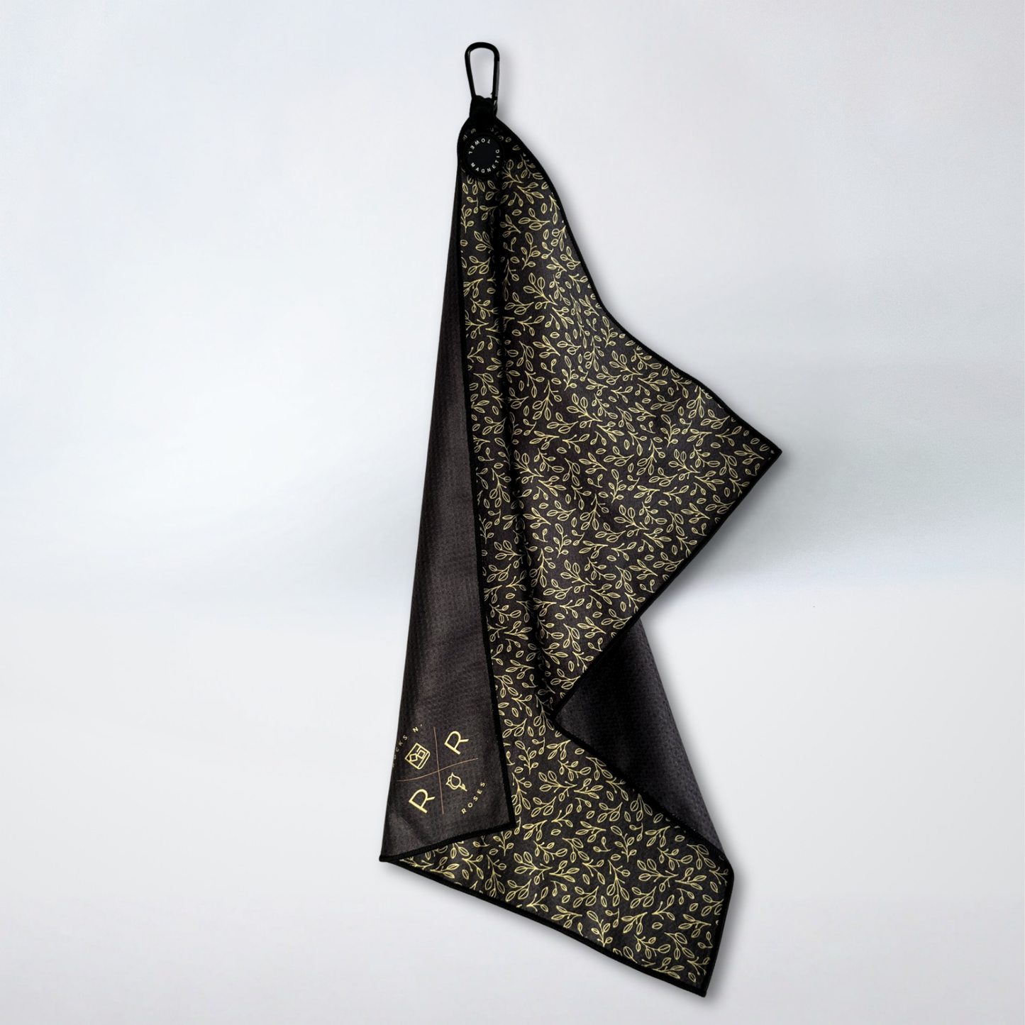 An image of the Rocks N' Roses Magnetic Golf Towel. On one side this magnetic golf towel has a black background with a golf floral pattern. The opposite side is plain black with a gold Rocks N' Roses logo in the bottom corner.