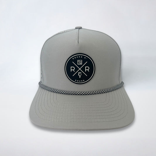 Image of the Rocks N' Roses 5 panel gray golf hat. This performance hat is made of 100% polyester, is water resistant, and has some stretch. It features a black and white circular rubber Rocks N' Roses logo on the front and a white and black diamond patterned rope.