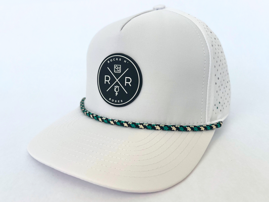 Close up shot of the Rocks N' Roses White Performance Golf Hat. This 100% polyester snapback hat is water resistant, offers some stretch and has that new school flat brim look. Also, it features a rubber logo and black, white, and teal rope that decorates the front brim. Laser cut venting for max breathability.