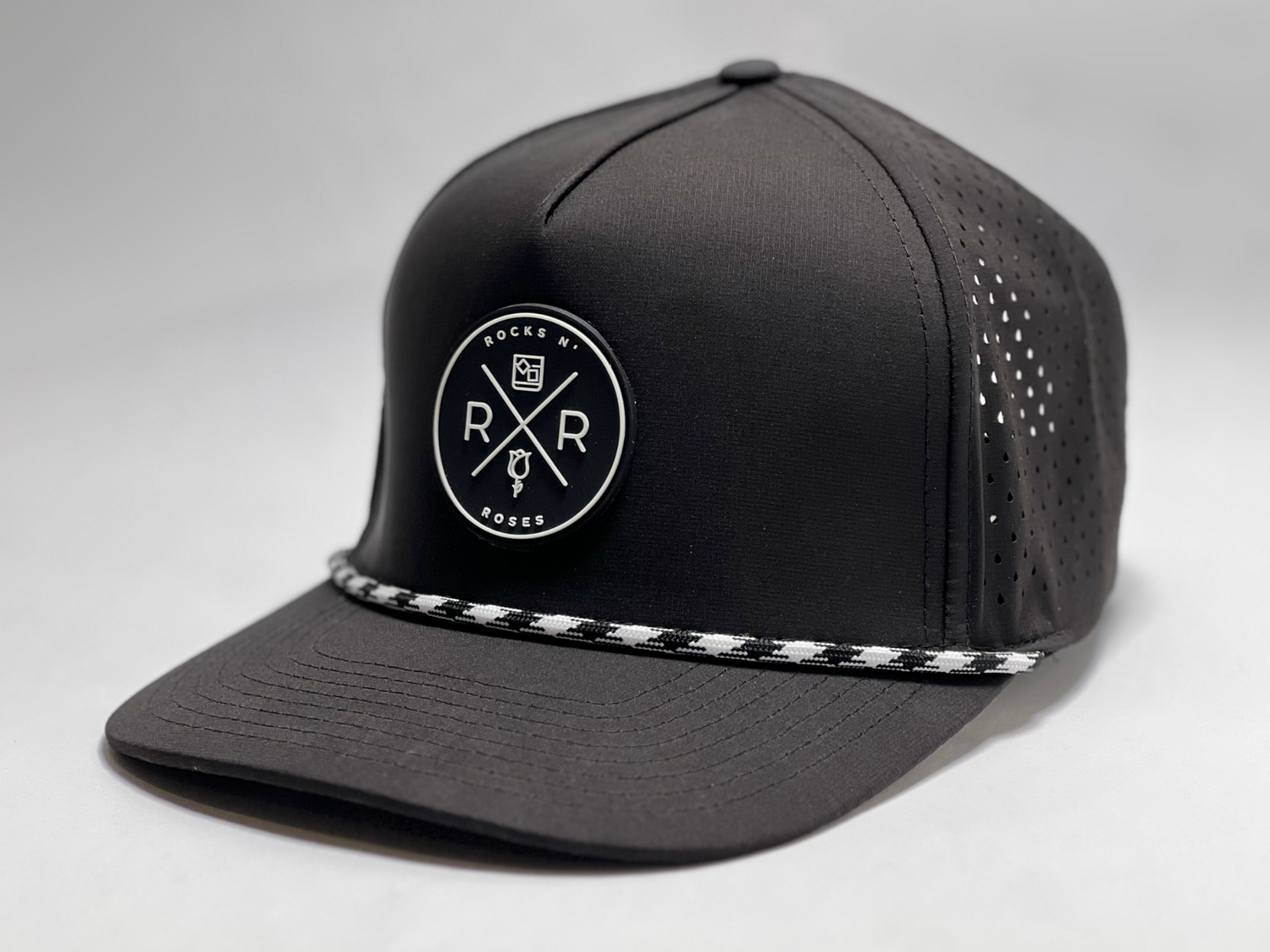 Close up shot of the Rocks N' Roses Black Performance Golf Hat. This 100% polyester snapback hat is water resistant, offers some stretch and has that new school flat brim look. Also, it features a rubber logo and black and white rope that decorates the front brim. Laser cut venting for max breathability.