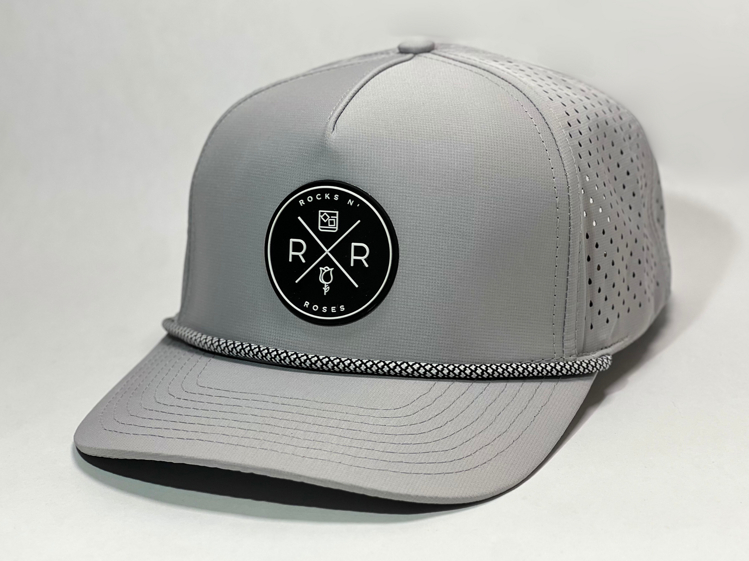 Close up shot of the Rocks N' Roses Gray Performance Golf Hat. This 100% polyester snapback hat is water resistant, offers some stretch and has that new school flat brim look. Also, it features a rubber logo and black and white rope that decorates the front brim. Laser cut venting for max breathability.