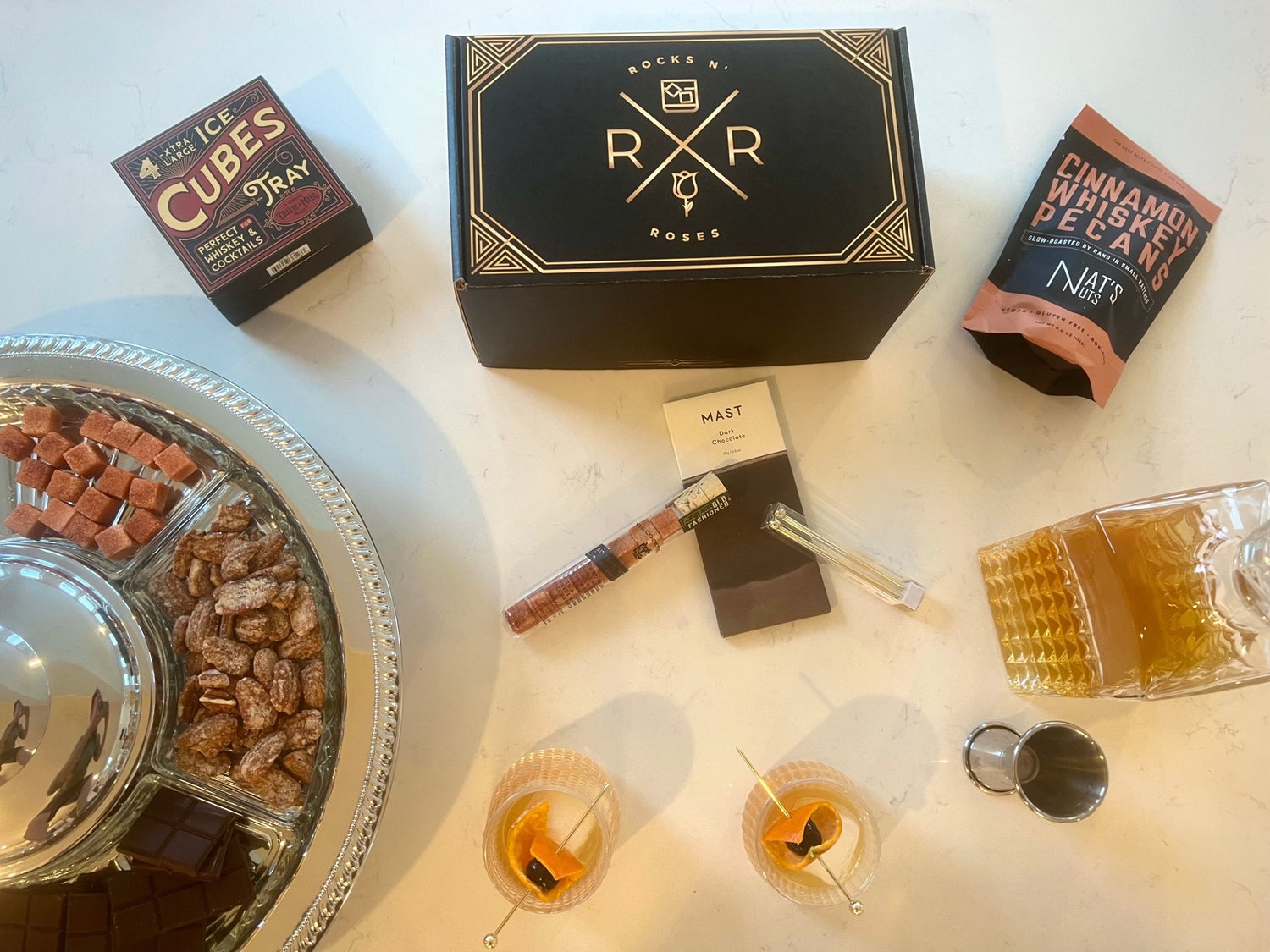 Overhead shot of the Rocks N' Roses Bourbon Box unpacked. Two Old Fashioned sitting on a table with a bottle of bourbon, cinnamon whiskey pecans, Old Fashioned sugar cubes, dark chocolate and two spinning whiskey glasses.