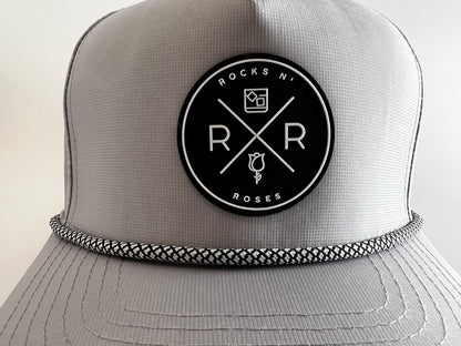 Close up photograph of the Rocks N' Roses gray performance golf hat made of 100% polyester. This closeup shows a circular rubber Rocks N' Roses logo in black and white and a black and white rope across the from of the brim.