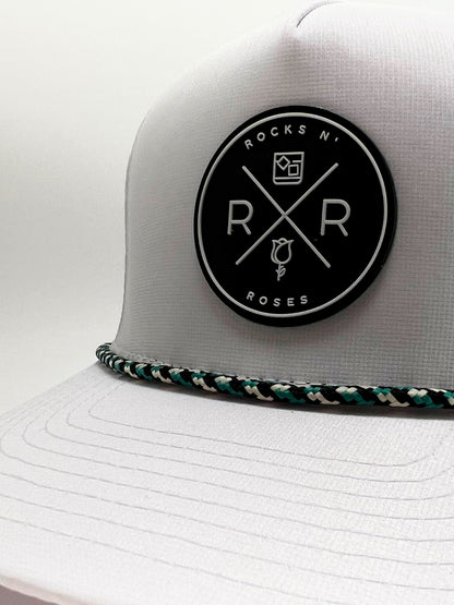 Close up photo showing the front of the Rocks N' Roses White Golf Hat which features a black rubber logo and white Rocks N' Roses brand logo. It also shows the performance hat's white, black, and teal rope.