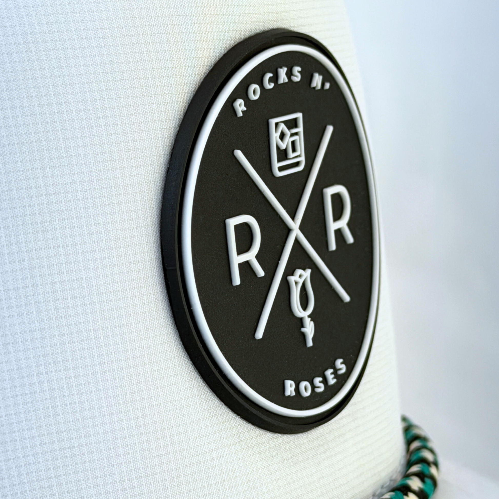 Close up photo showing the front of the Rocks N' Roses White Golf Hat which features a black rubber logo and white Rocks N' Roses brand logo. It also shows the performance hat's white, black, and teal rope.
