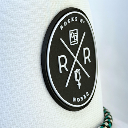 Close up photo showing the front of the Rocks N' Roses White Golf Hat which features a black rubber logo and white Rocks N' Roses brand logo. It also shows the performance hat's white, black, and teal rope.