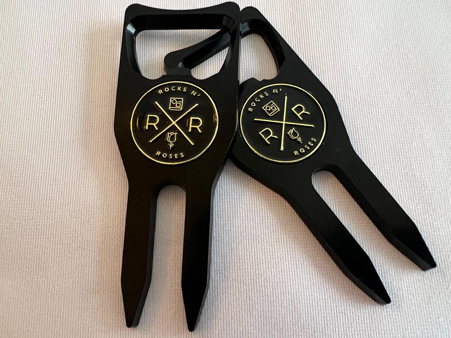 Image of two Rocks N' Roses black and gold golf multi-tools. These golf multi-tools combine a divot repair tool with a magnetic ball marker, bottle opener, and club rest.