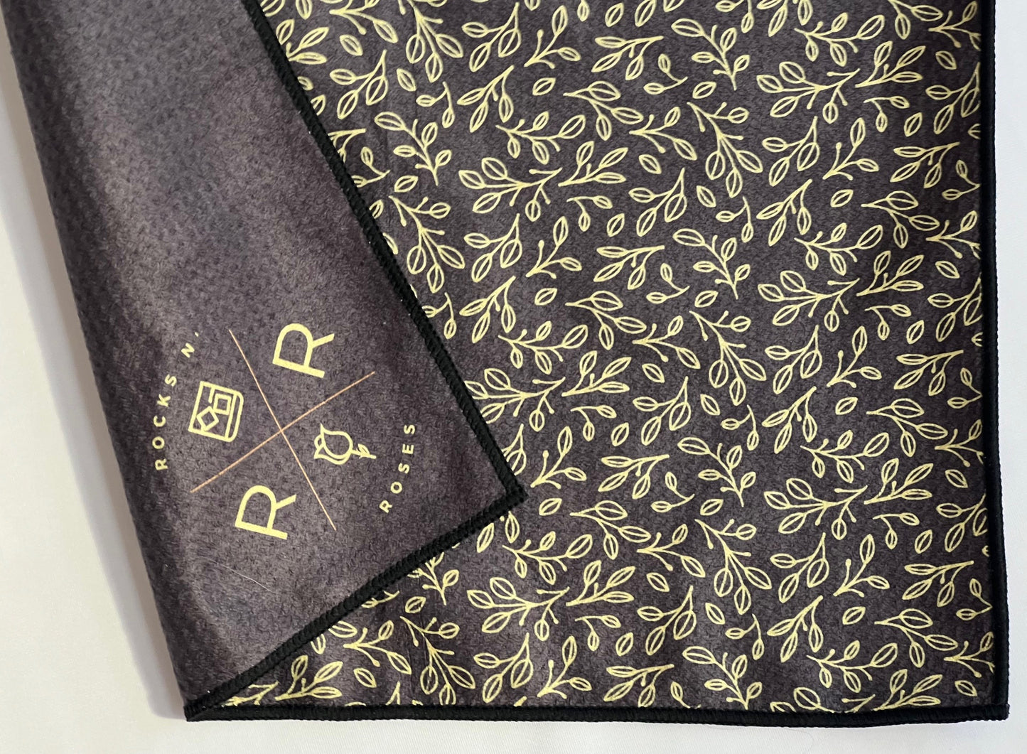 A close up shot of Rocks N' Roses' black and gold floral pattern magnetic golf towel featuring a waffle weave synthetic material and gold Rocks N' Roses logo.