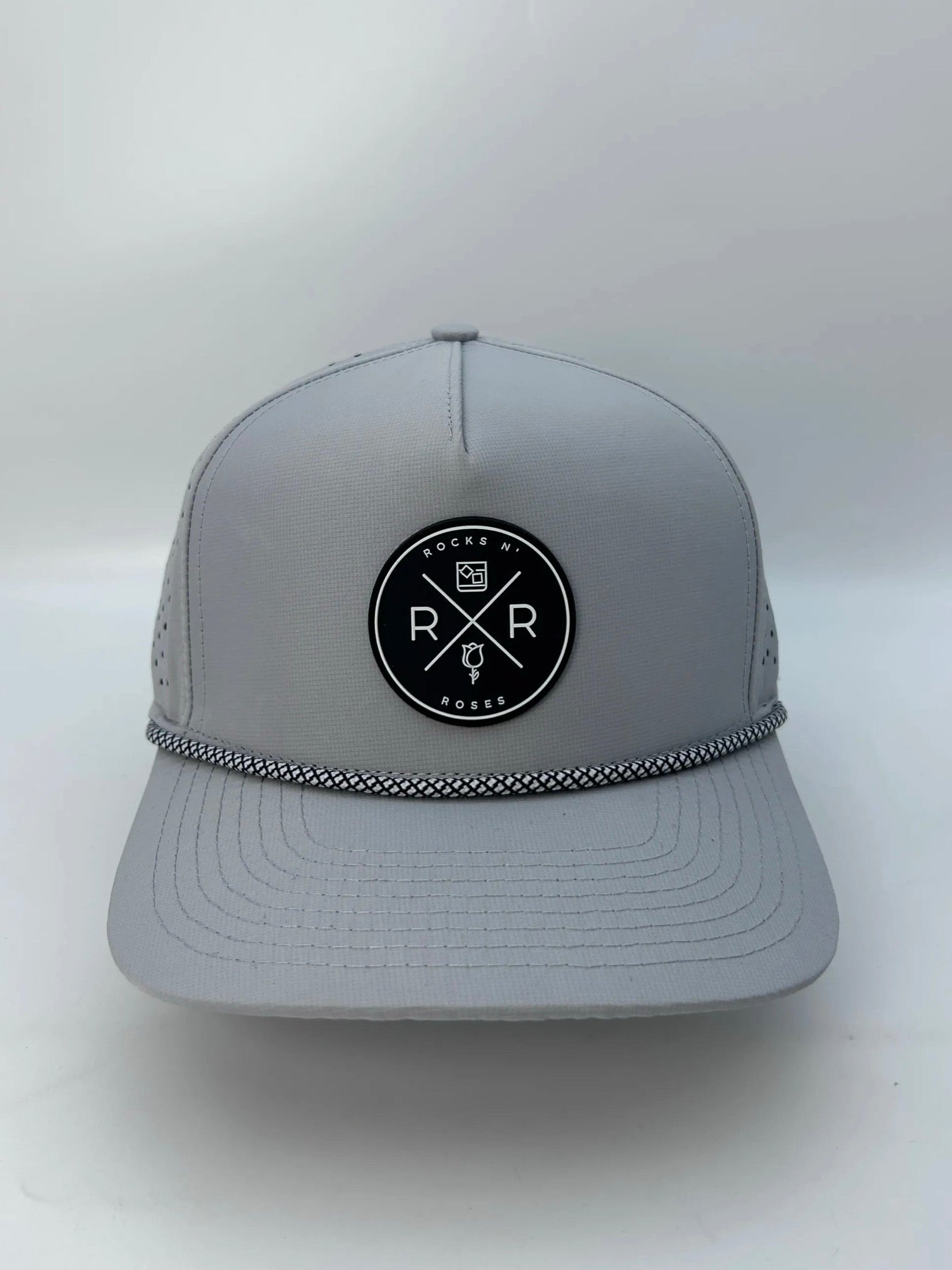 Image of the Rocks N' Roses 5 panel gray golf hat. This performance hat is made of 100% polyester, is water resistant, and has some stretch. It features a black and white circular rubber Rocks N' Roses logo on the front and a white and black diamond patterned rope.