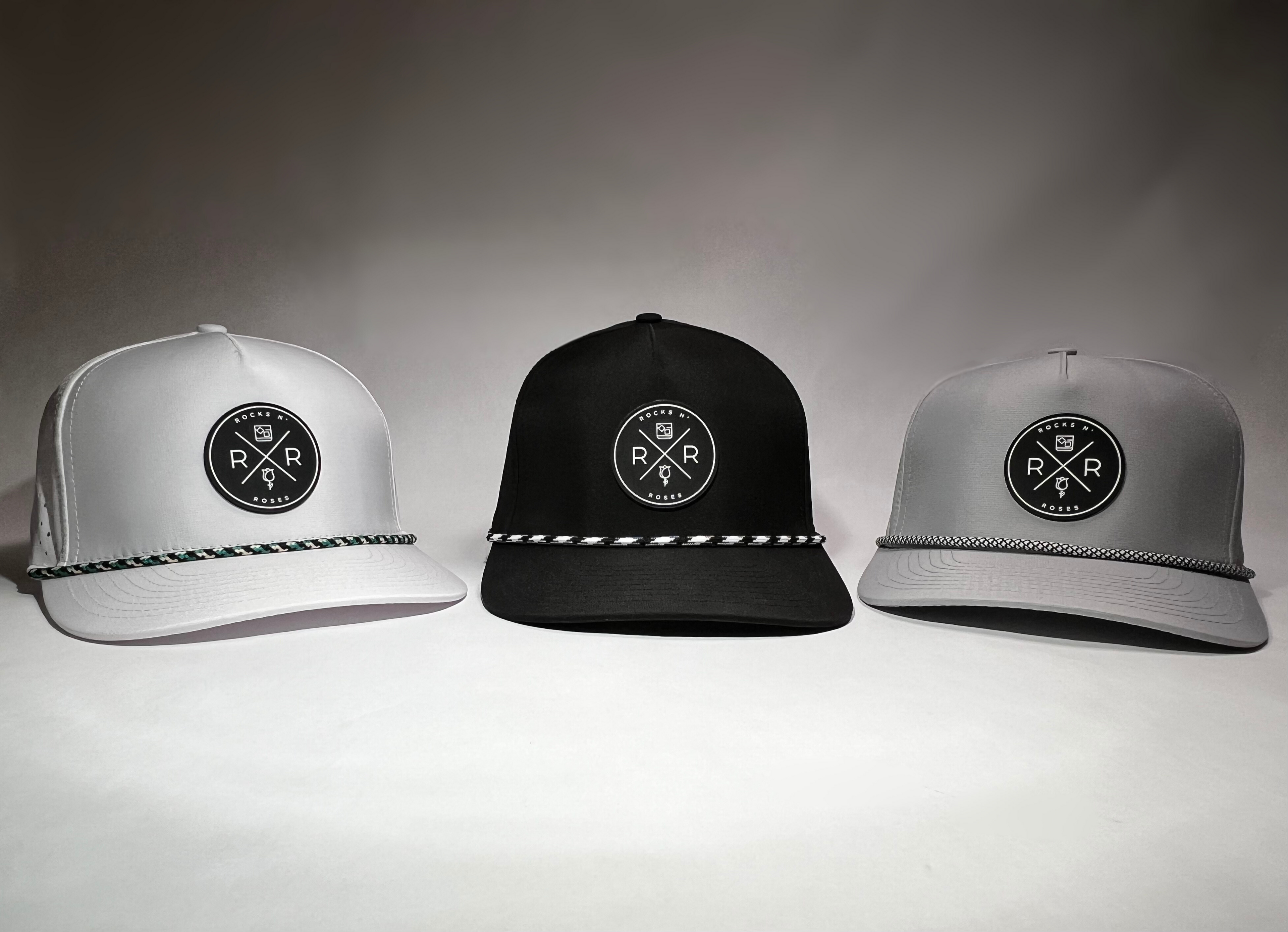 Image of the Rocks N' Roses Performance Golf Hat Collection feature a white, black and gray golf hat. Each hat features a circular rubber Rocks N' Roses logo and rope on the brim.