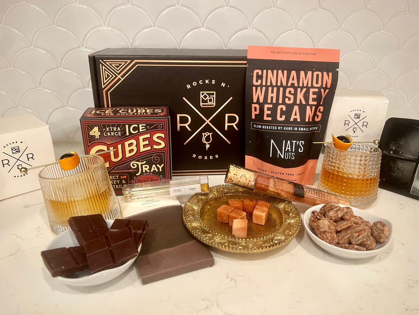 An image of the Rocks N' Roses Bourbon Box unpacked. Featuring two spinning whiskey glasses, Yes Cocktail Co.'s five spice organic Old Fashioned sugar cubes, Nat's Nuts cinnamon whiskey pecans, Trixie & Milo's BPA Free xtra large ice cubes, Mast Brother's dark chocolate bar and Luxe's cocktail spears.